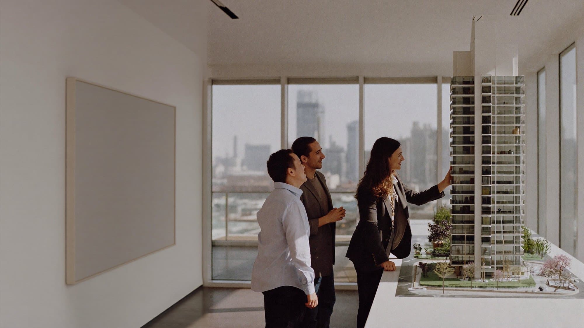 A sales agent walking a couple through a scale model of a residential tower in a bright gallery